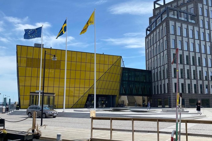 The photo shows an impression of the Linneaus campus in Kalmar. In the foreground is a wooden walkway and, farther back, a paved square. Behind it, in the centre of the picture, is a modern, geometric building in yellow, in front of which the flags of the university and of Sweden and the EU are flying.  To the right is another building. The sky above the scene is light blue with white feather clouds. 