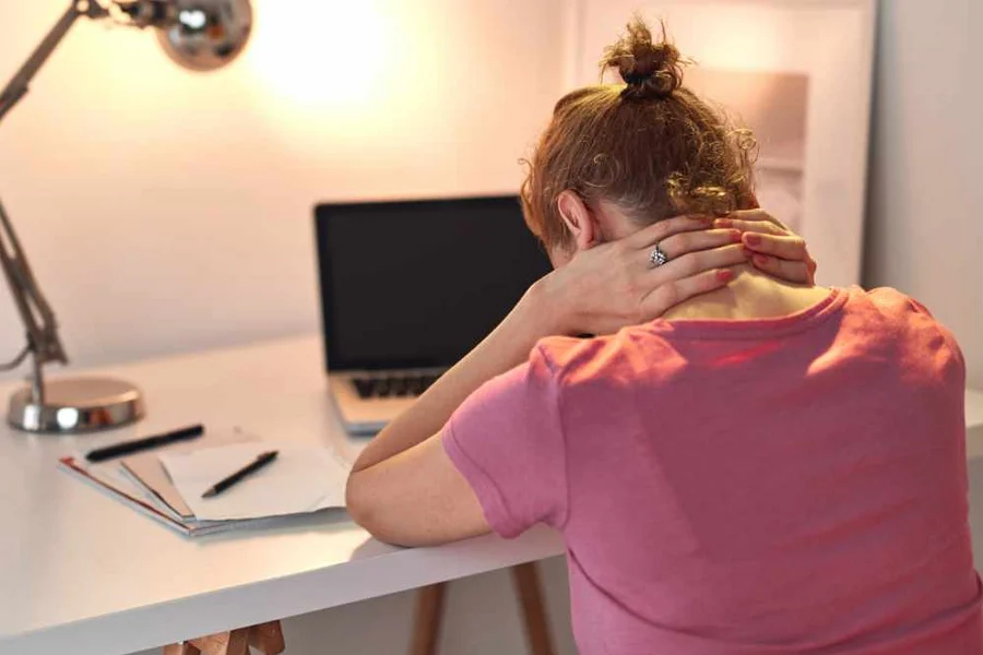 Woman in a pink T-shirt sitting at a desk with a laptop and notebooks, holding the back of her neck with both hands, suggesting neck pain or tension.