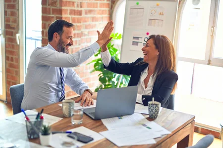 A picture of a man and a woman sitting at a table in front of a laptop in a bright room, high-fiving each other.