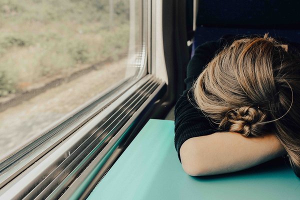 The photo shows a person on a train. All that can be seen of her head is the blond hair tied into a bun, which rests on her arm on a small green train table. On the left of the picture is a section of the window with a passing piece of meadow. 