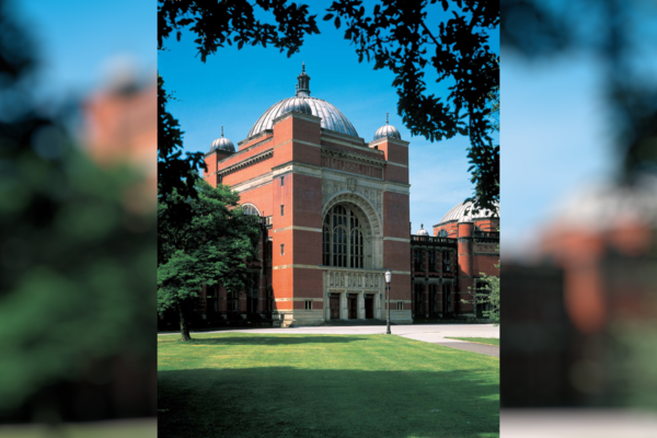 The photo shows the main entrance to the University of Birmingham, a magnificent red brick building with a domed roof, a green lawn in front. The picture is framed by branches of the surrounding trees.
