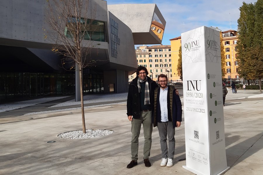 The photograph shows two men in front of the MAXXI (National Museum of XXI Century Arts) in Rome.