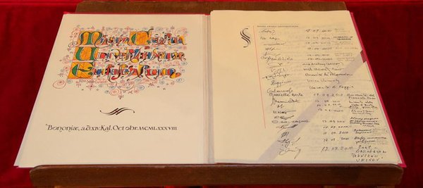A photo of the original Magna Charta document. On the left-hand side, "Magna Carta Universitatum" can be read in ornate lettering with the place and date of the signing “Bologna, October 1988” written below; the right-hand side shows a series of signatures.