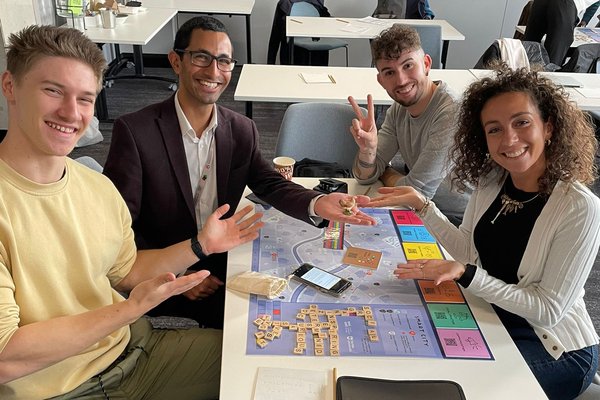 Four enthusiastic young participants engaging with a colourful board game, smiling and posing with hand gestures, in a bright office setting.