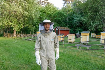 The photo shows a young man, Oscar Bergström, in Beekeeper attire on a green meadow. In the background are numerous bee boxes, with a row of trees behind them.