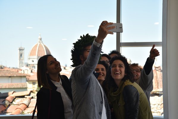 The photo shows several students standing close together, in their midst one person has raised their arm with a mobile phone to take a selfie of the group. The dome of the Florence Cathedral can be seen in the background.