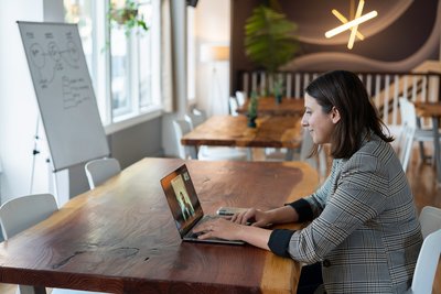 The photo shows a young woman at a laptop where she is participating in a video conference.  She is sitting at a wooden table, in the background are other identical tables and a flipchart.