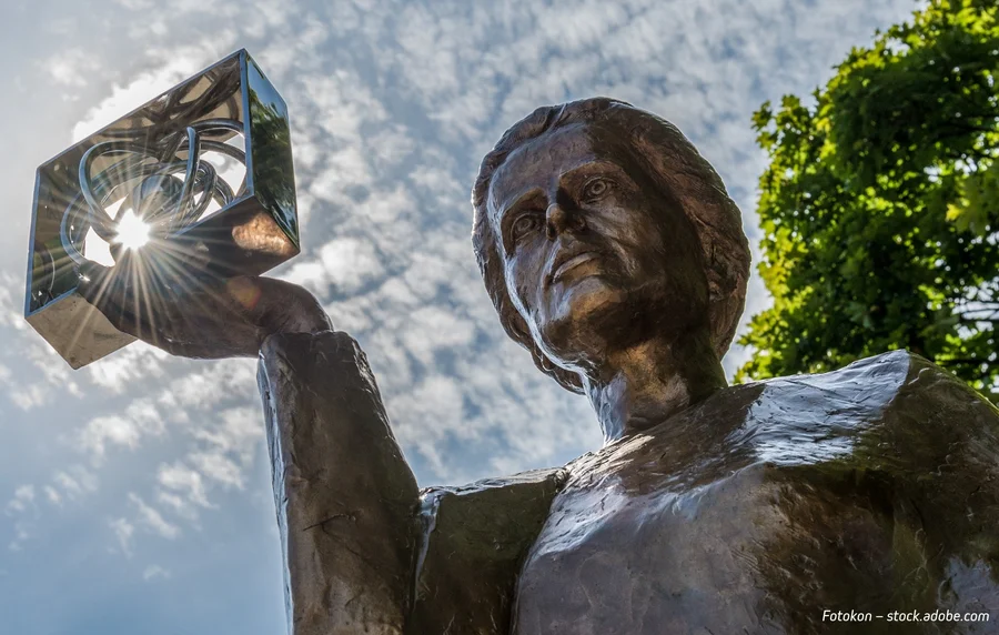 Bronze statue of Marie Curie holding a glass cube, viewed from below against a blue sky and trees.