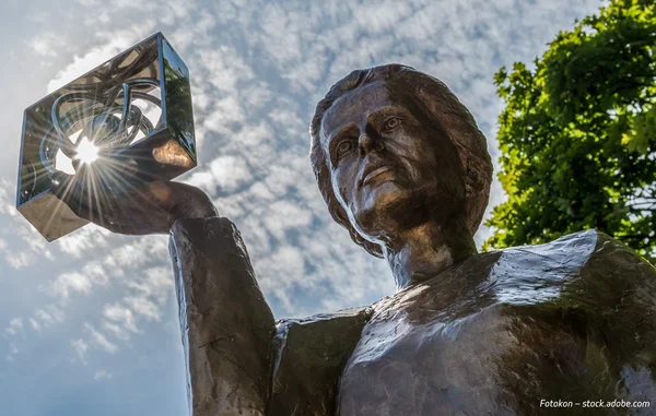 Bronze statue of Marie Curie holding a glass cube, viewed from below against a blue sky and trees.