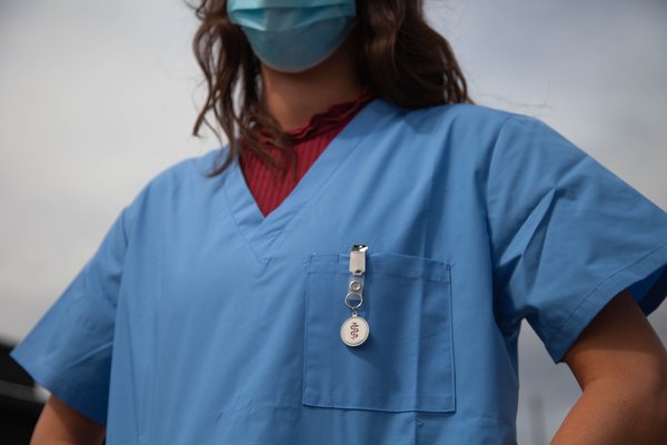 The photo shows a woman with brown hair wearing a light blue nurse's uniform over a long-sleeved red top. She has her hands on her hips. Only the lower half of her face is visible, which is covered by a medical mask.