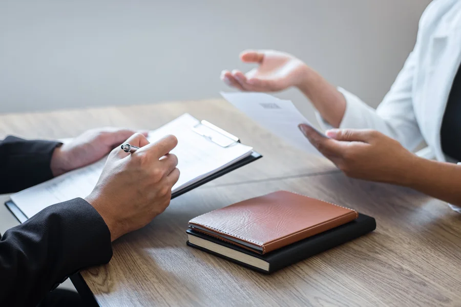 A close-up of four hands on a desk. One person is writing notes in a notebook, while the other is holding a piece of paper. The desk also has two notebooks, one brown and one black..