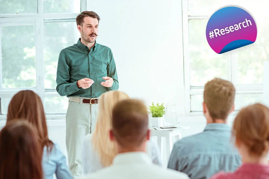 This image shows a person giving a presentation to a group. There is a colourful bubble with the hashtag #Research visible, suggesting that the course is related to the EUniWell #Research project. The atmosphere appears to be professional and educational.