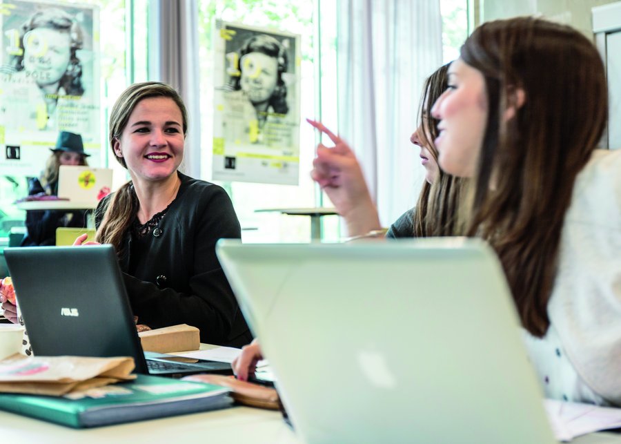 The picture shows three female students who are sitting at a table with their laptops and are in the middle of a conversation.