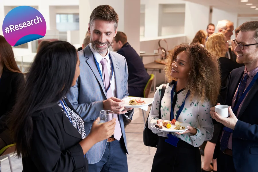 The image depicts a professional networking event where a group of people are engaged in conversation. They appear to be enjoying their interactions with each other, some holding beverages and plates of food. A noticeable feature is the #Research sign. The individuals are dressed in business attire, indicating a formal or semi-formal gathering.