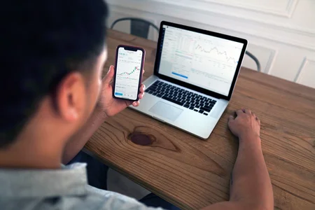 A man looking at a smartphone and a laptop, both showing graphs on their screens