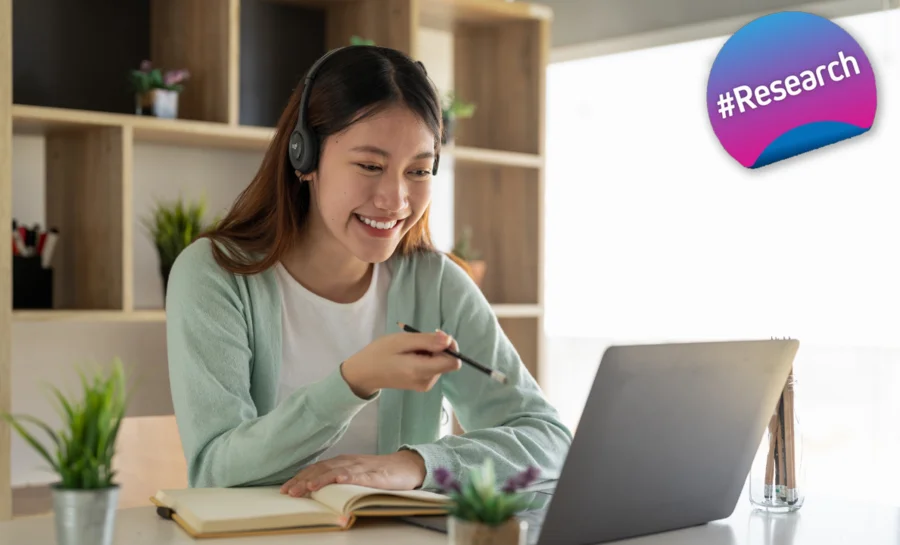 A young woman sits at a desk, a notebook and an open laptop in front of her. She is wearing a headset and smiling towards the screen. In the top right-hand corner is a round graphic on which the word #Research can be read on a gradient background from blue to pink.