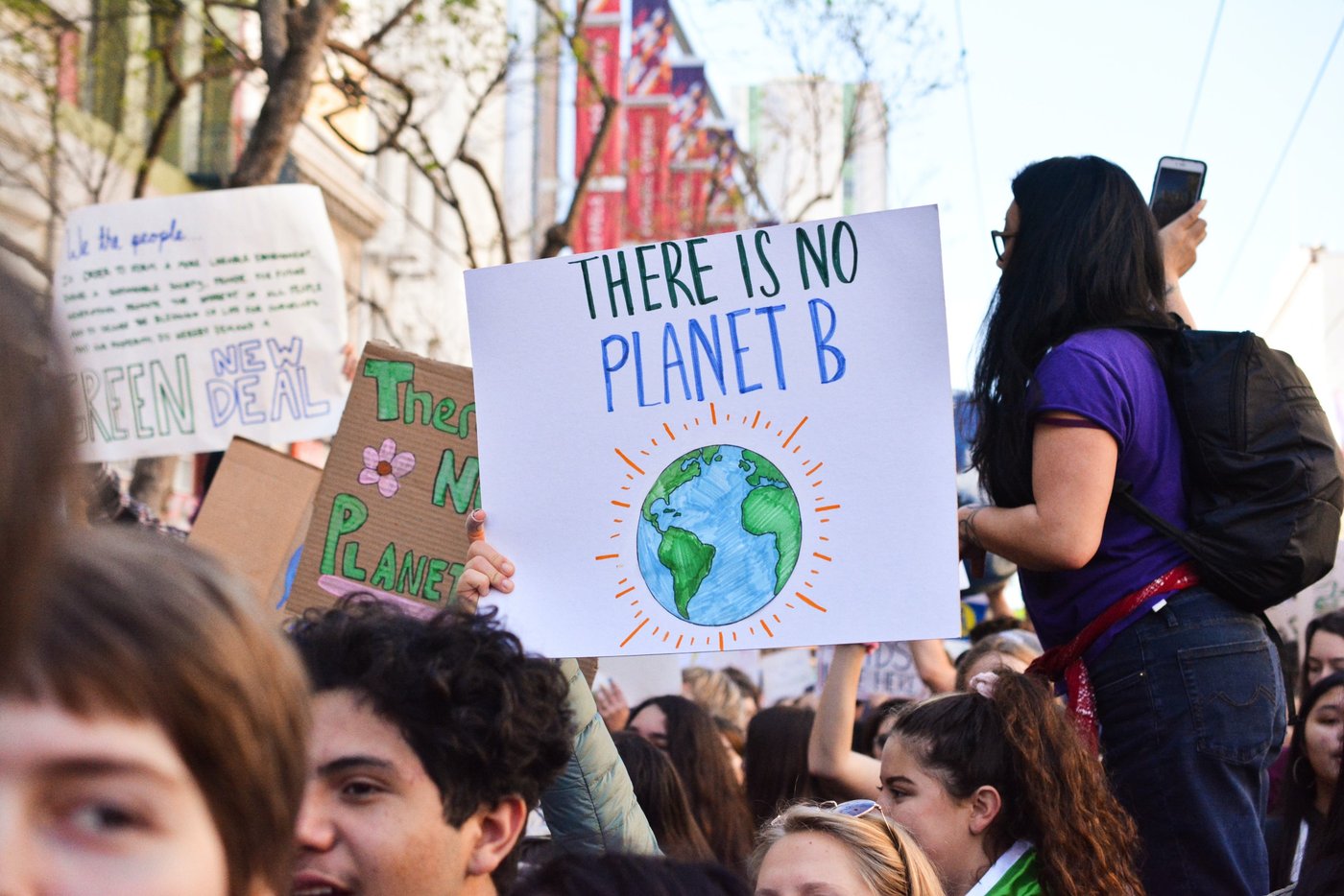 The photo shows young people at a demonstration. In the centre is a sign that says "There is no planet B" with an illustration of planet Earth underneath. 