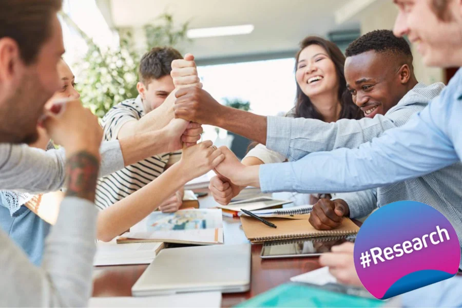 The image shows a group of individuals in a collaborative setting, engaging enthusiastically with each other. They appear to be celebrating a success or breakthrough, in a research or educational context, showcase by the hashtag #Research badge displayed in the lower right corner.
