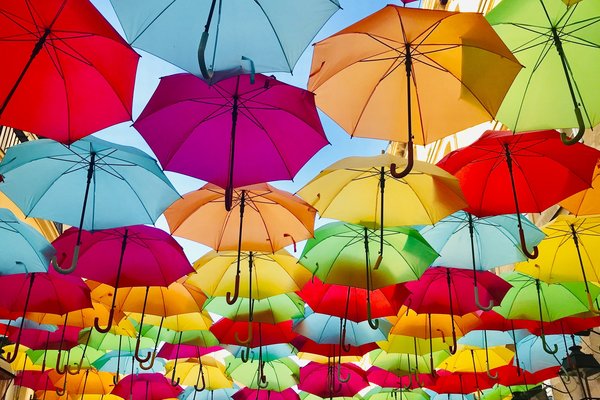 Colourful umbrellas are suspended between buildings under a blud sky in the background.