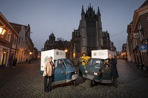Three people are standing on a paved square surrounded by buildings, with a church in the background. They are standing at the open doors of two three-wheeled vans bearing the logo of the European City of Science Leiden 2022. The persons are, from left to right: Annetje Ottow, Auke-Florian Hiemstra, and Leiden Mayor, Henri Lenferink.