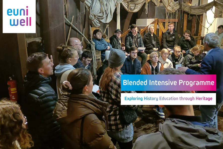 A group of students gathered in a museum, listening to a guided tour. In the top left corner, the 'EUniWell' logo is visible, while the center of the image displays the course title: 'Blended Intensive Programme - Exploring History Education through Heritage'