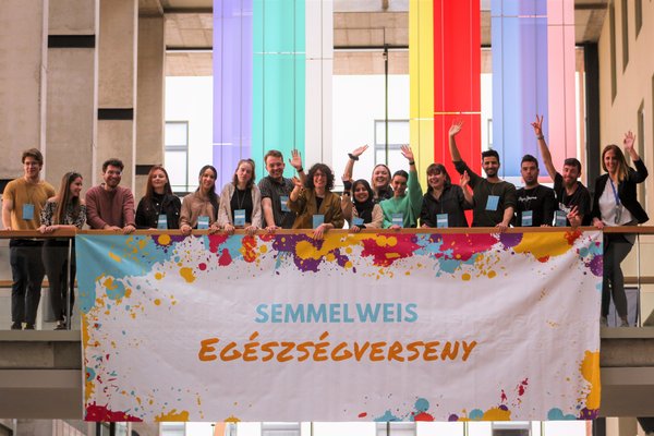 A group of students wave from a passageway. A banner with the logo of the Semmelweis Health Competition is attached to it. Colourful flags hang in the background.