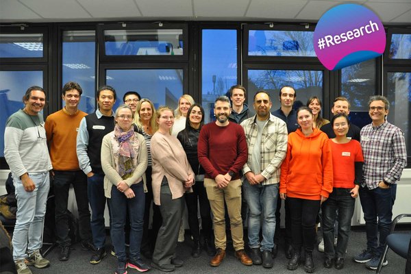 A group of seventeen individuals are gathered in a room with a "#Research" sign, smiling for a team photo. They are dressed in casual and smart-casual attire, with a diverse mix of ages and backgrounds. The setting is indoors with natural light from windows.