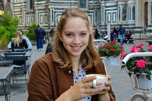 The picture shows Lotte Nagelhout, holding a cup of coffee and smiling at a sidewalk coffee shop.