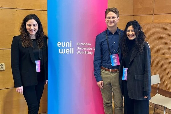 A picture of the EUniWell Student Board Executive team (Sloan Kudrinko, Liam Walsh, Letizia di Donato) in front of a EUniWell flag.