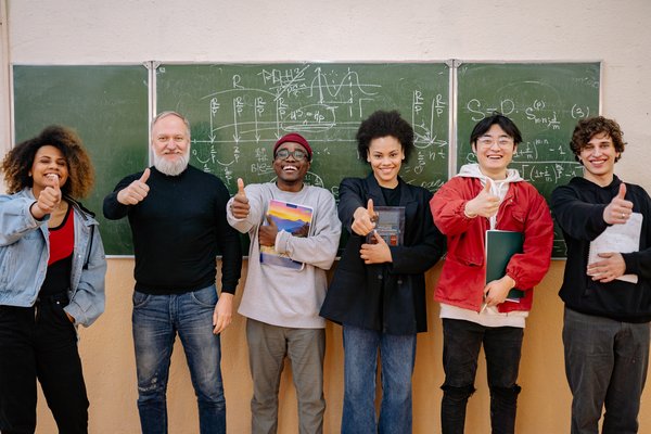 Six people standing in front of a chalkboard, smiling with their thumbs up.