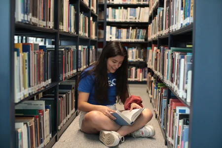 A student sitting on the floor of a library, surrounded by bookshelves.