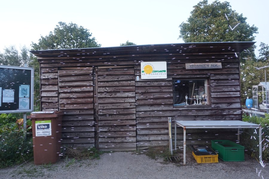 A rustic wooden shed with weathered planks stands surrounded by greenery. A sign reading 'HET ZONNEVELD' is affixed to the shed, alongside a window displaying various items. Nearby, an informational panel and a brown bin labelled 'Vilko' are placed. Beneath the window is a metal table sheltering a couple of storage bins.