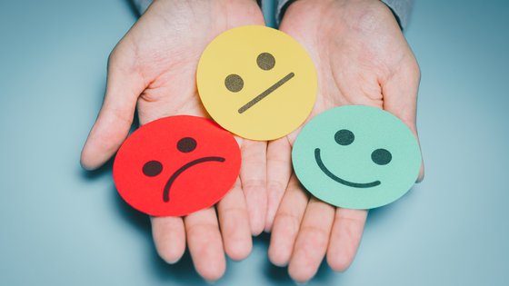 Photo of hands holding 3 cardboard faces: 1 red sad, 1 yellow straight, 1 green happy