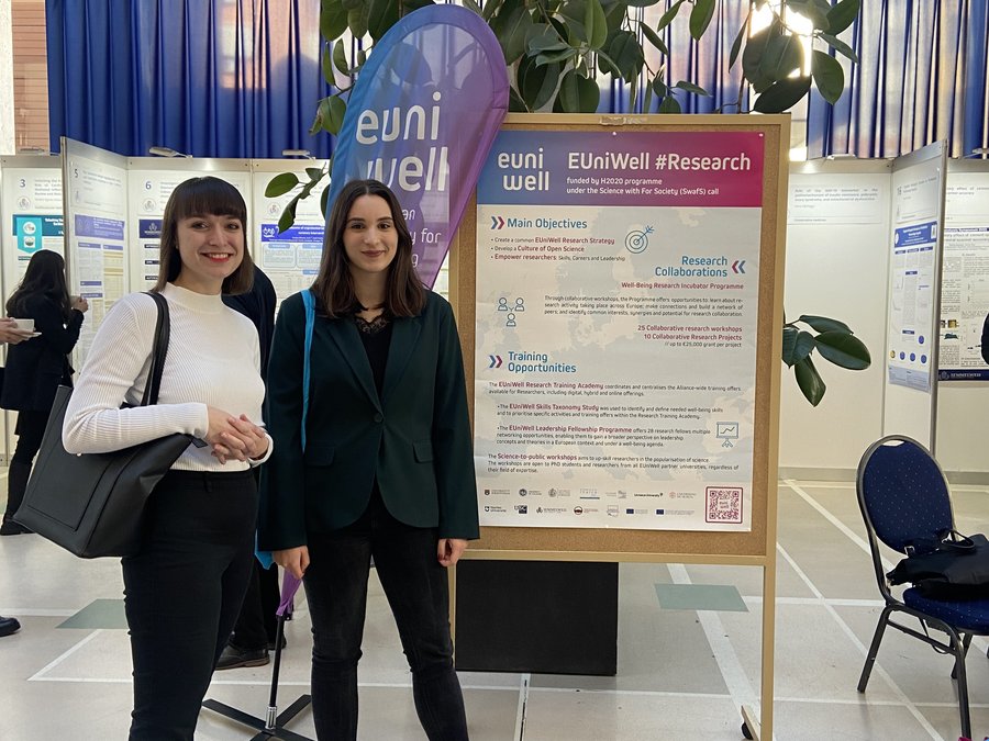 Two individuals are posing in front of a EUniWell information banner detailing research objectives, collaborations, and training opportunities at a university event.