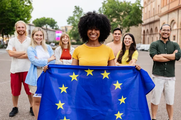 Seven smiling young adults stand outdoors, with a woman at the centre holding a European Union flag in the foreground.