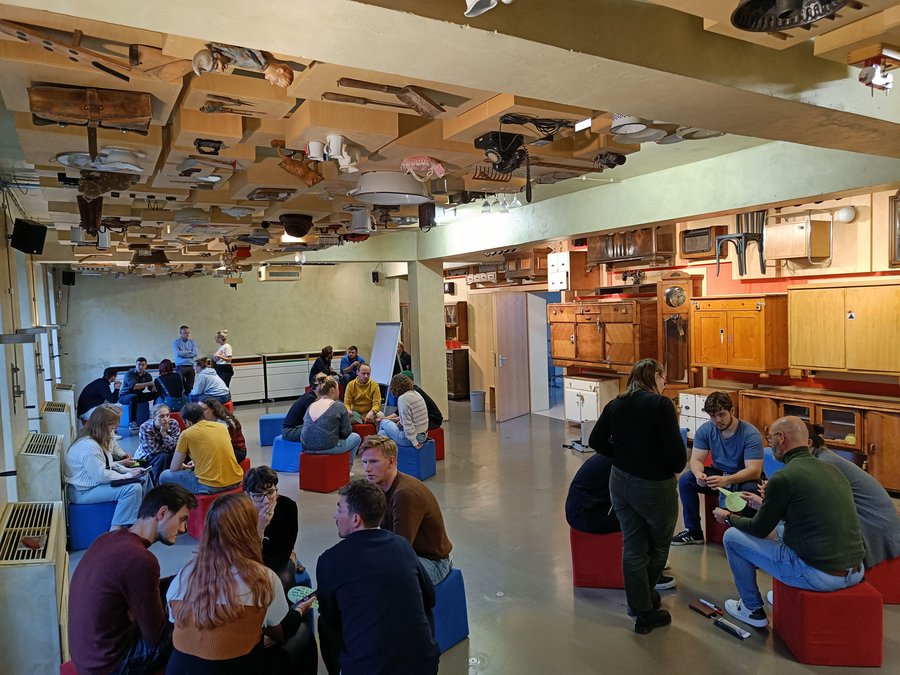 Students sitting in different groups in a museum space. The wall and ceiling are decorated with vintage furniture and everyday objects.