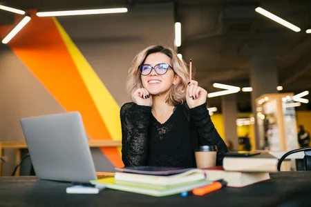 A person sitting at a table with a laptop, books, and a coffee cup, holding a pencil with the hand and with a expression of thinking on ideas