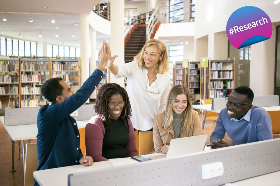 A group of 5 young professionals in a library. 2 persons are hive-fiving, while the others are looking at a laptop, smiling. In the upper right corner is a badge with the text "#Research".