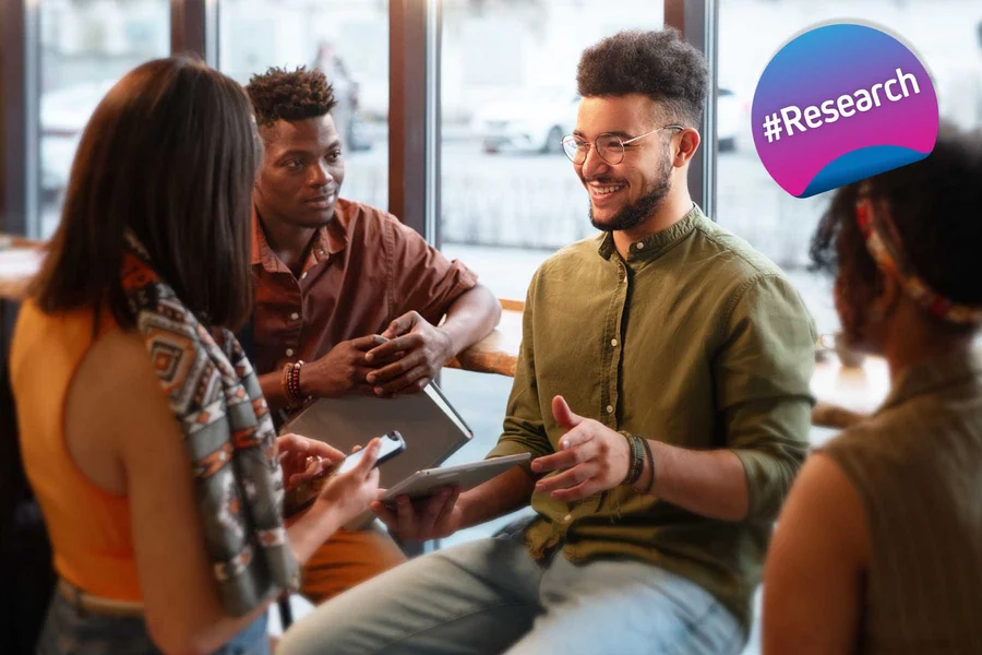 A group of individuals in a collaborative setting, with one person holding a digital tablet. In the upper right corner, there's a digitally added badge with the hashtag #Research.