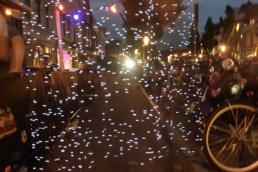 Blurry night-time scene of a city street, illuminated by streaks of white lights, with bicycles parked on the side and a glimpse of buildings and street lamps in the background.