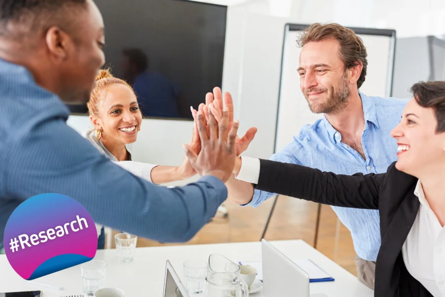  The image depicts a group of four professionals in a meeting room engaging in a teamwork gesture. Two individuals are high-fiving each other, signifying a successful collaboration or agreement. The other two are smiling and appear to be part of the celebration. The atmosphere is positive, emphasising a sense of accomplishment and mutual support. In the foreground, there’s a badge with the hashtag #Research, indicating that the picture is related to the EUniWell #Research project.