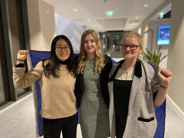 A photo of three representatives of the EUniWell Student Board. Cho Park, Rebecca Esselgren and Judith Barth are standing next to each other in a hallway with an EU flag draped over their shoulders.