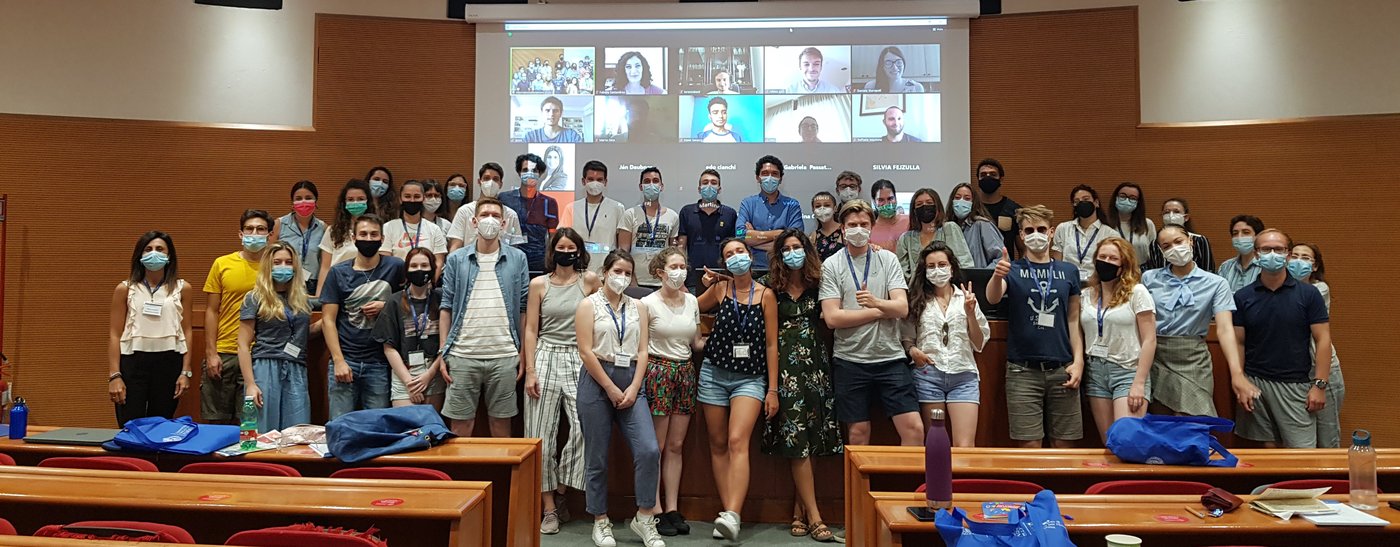 The photo shows a group of students in a lecture hall, all wearing masks. In the background is a screen showing the digital participants. 