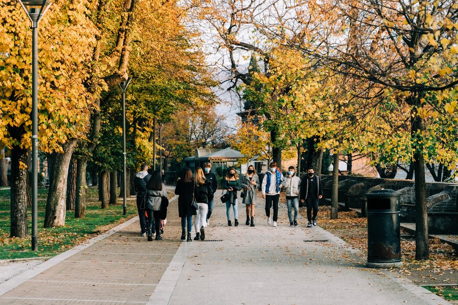 The photo shows a group of young people wearing medical masks alongside everyday clothes. They are walking towards the camera on a paved path, with autumnal trees to the right and left along the way.