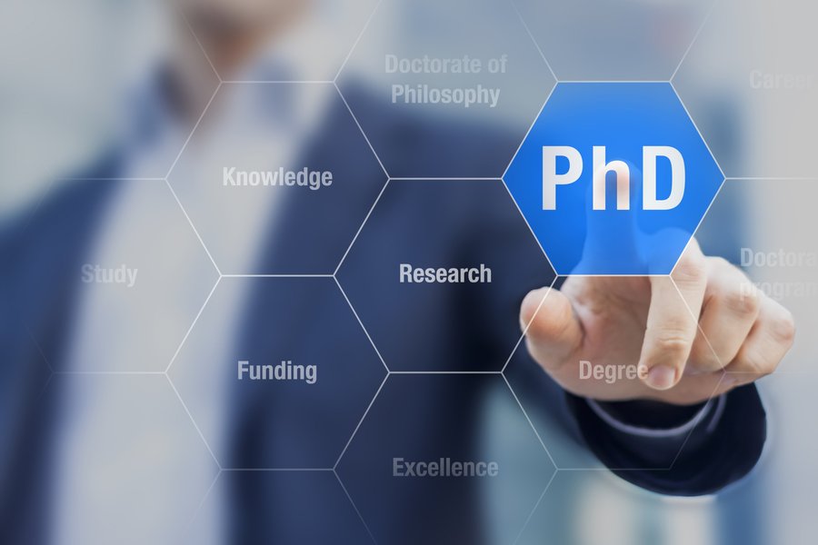 The picture shows a person, blurred in the background, pointing with his finger at a tile with the word PhD on it. 