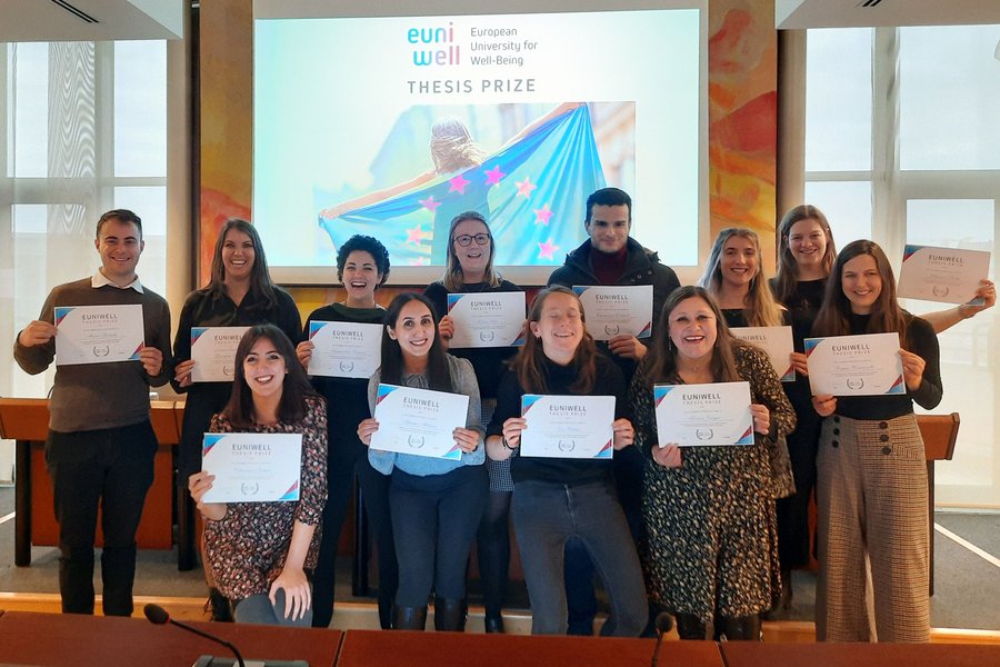 The picture shows the recipients of the EUniWell Research Thesis Prize being honoured. They are holding up a certificate, behind them the EUniWell logo below which it says "Thesis Prize" and a picture of a girl holding an EU flag is projected, the stars appearing in EUniWell magenta.