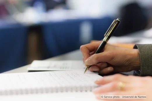 The photo shows the close-up of a hand writing on a sheet of paper lying on a desk.