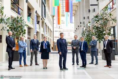 Nine representatives of Semelweis University pose in an entrance hall, plants stand to their right and left, colourful flags hang from the ceiling, a large window can be seen in the background.