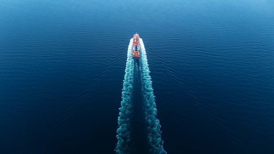 Wide-angle photo of a cargo-ship cutting through the sea with a tail of foam