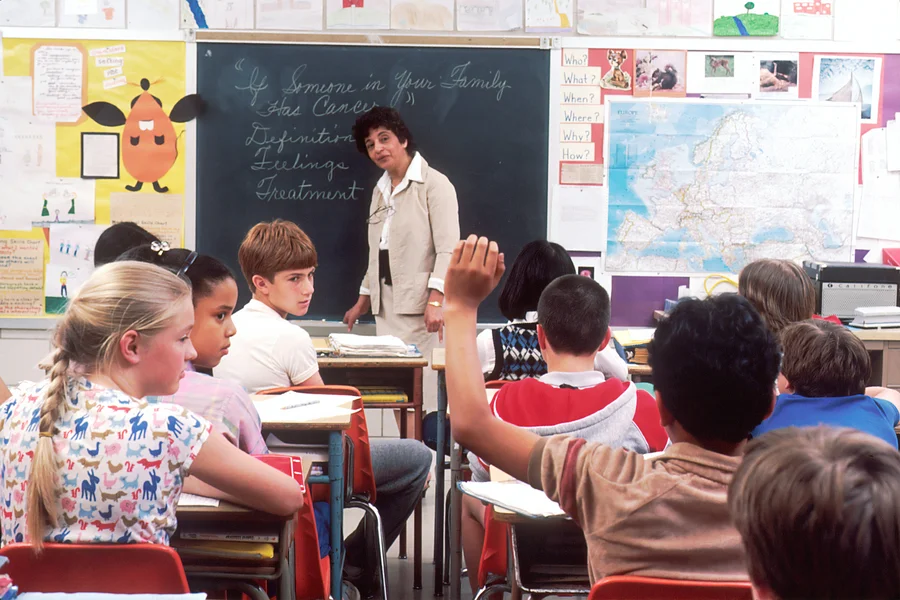 The photo shows a classroom with pupils, a teacher is standing at the blackboard talking to the class.